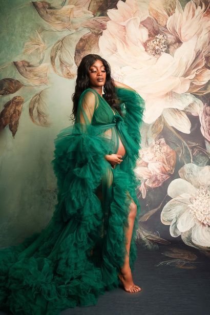 A pregnant woman in a green dress is standing in front of a floral wall