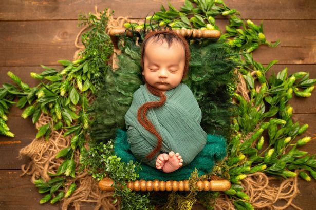 A newborn baby wrapped in a green blanket is sleeping in a wooden crib surrounded by greenery