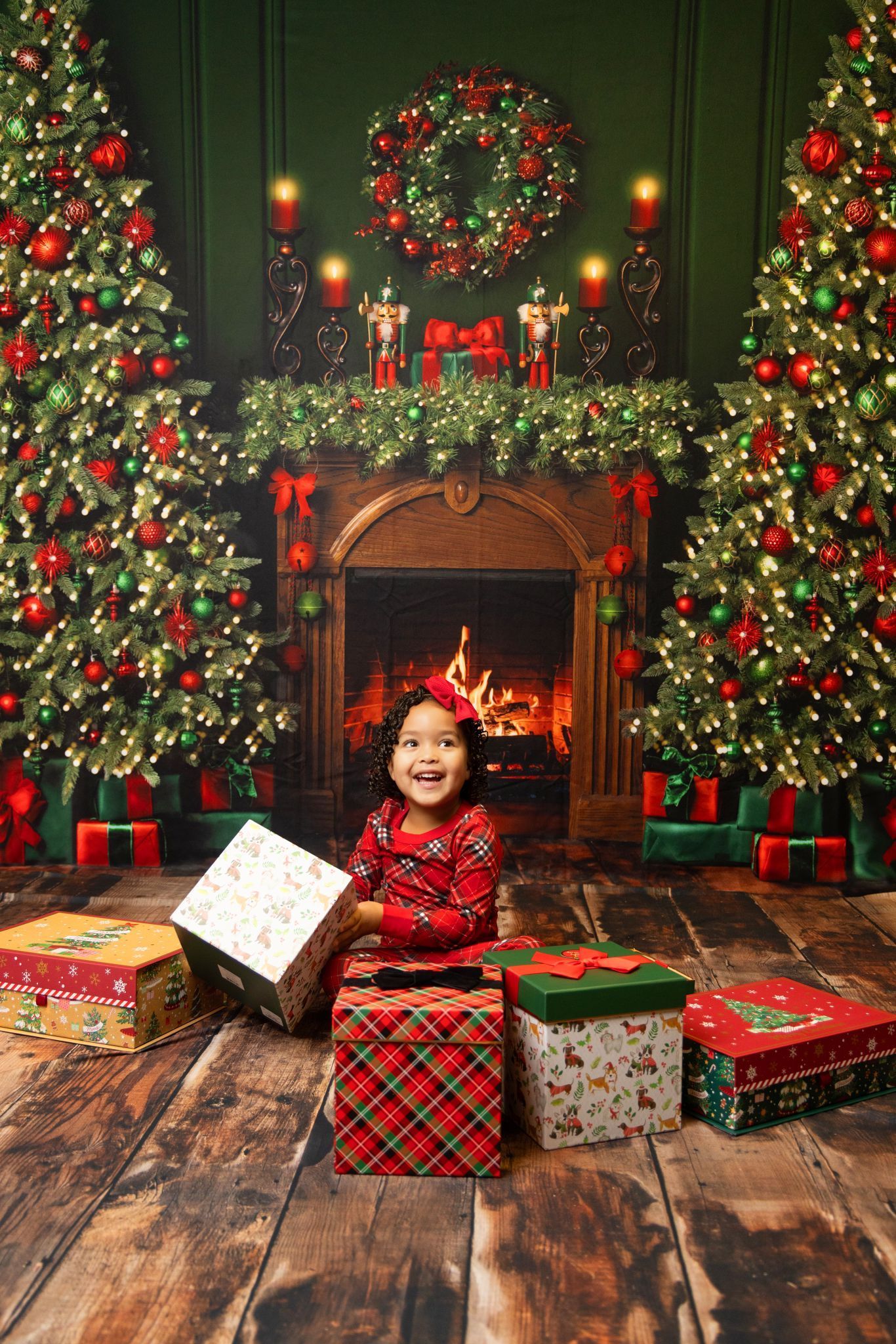 A little girl is sitting in front of a fireplace with christmas trees and presents.