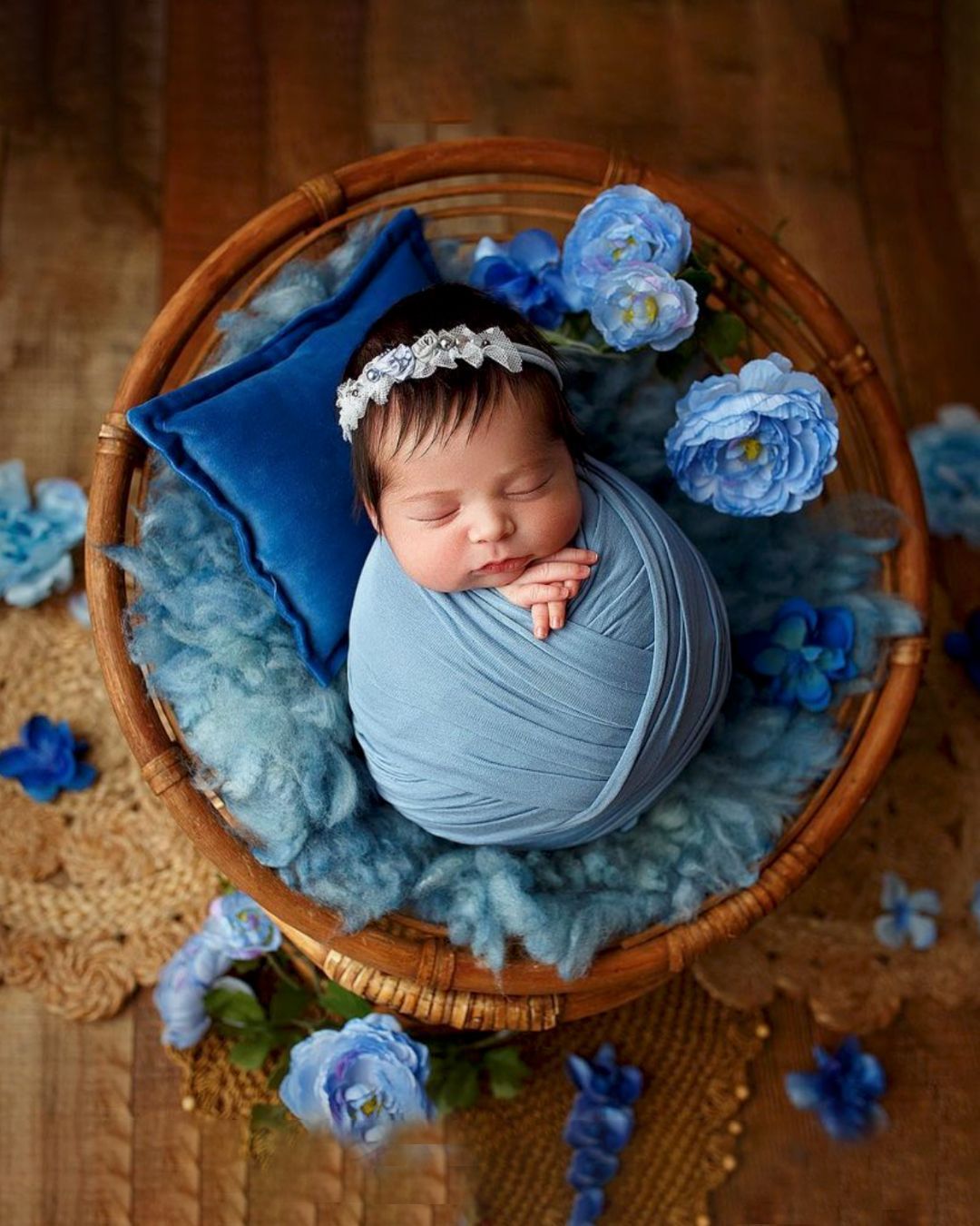 A newborn baby wrapped in a blue blanket is sleeping in a wicker basket surrounded by blue flowers