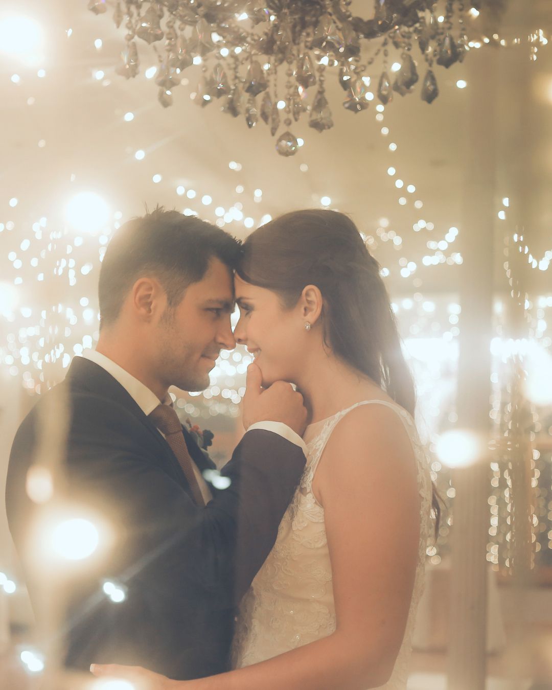 A bride and groom are touching their foreheads in front of a chandelier