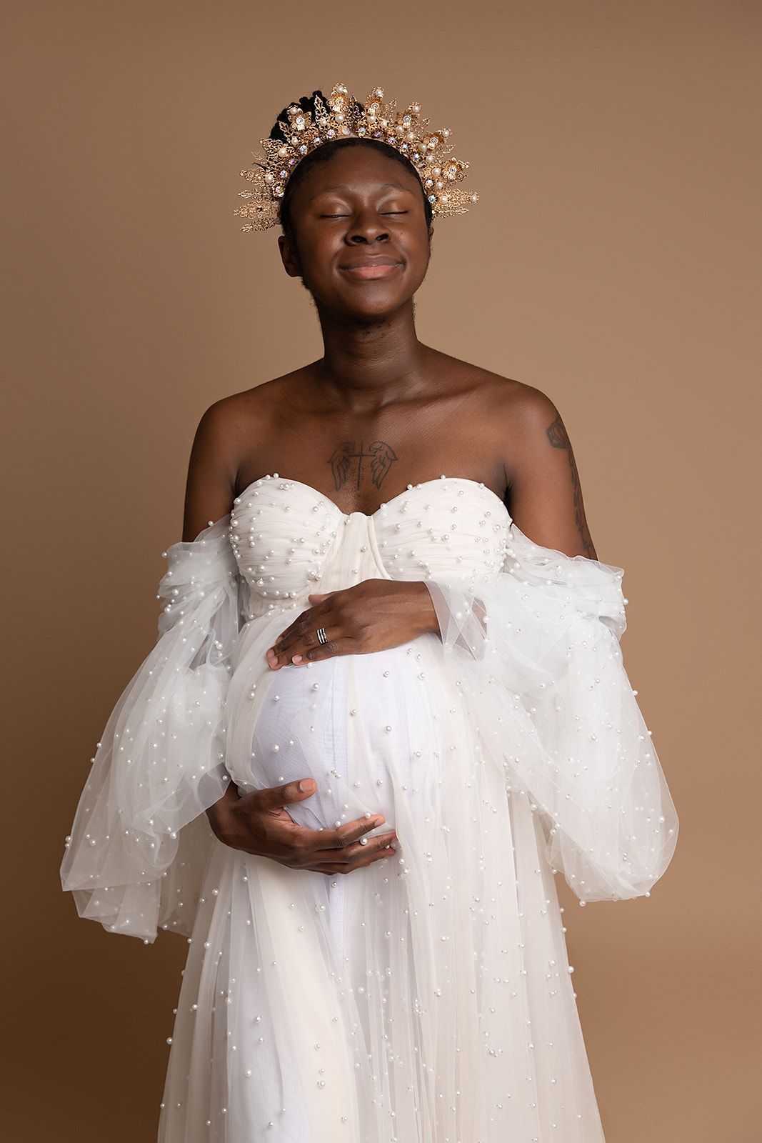 A pregnant woman in a white dress and crown is holding her belly