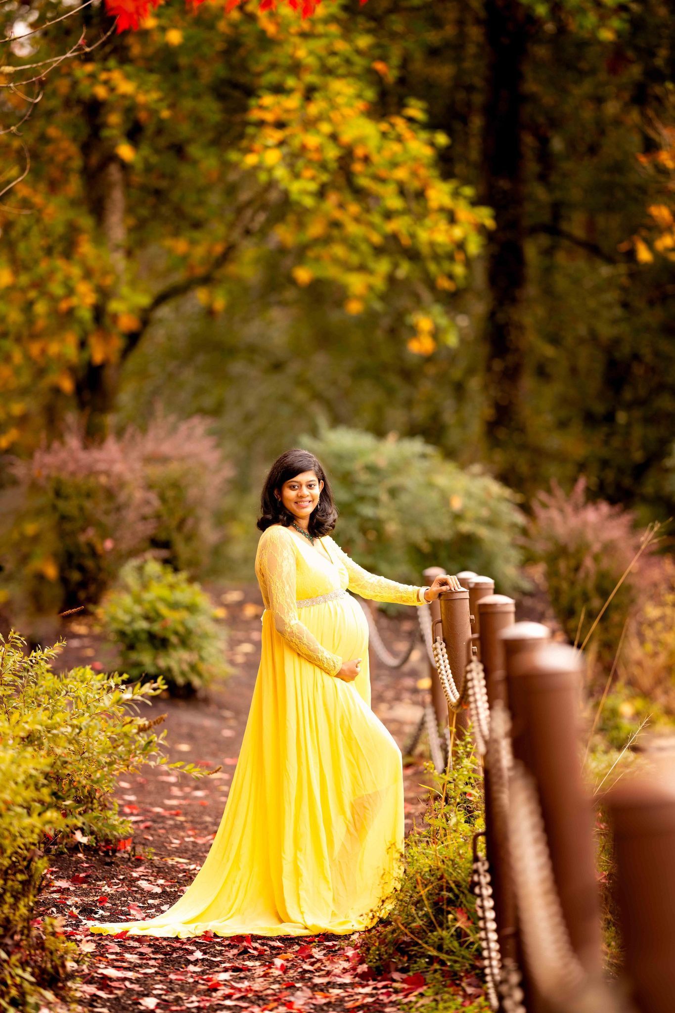 A pregnant woman in a yellow dress is standing next to a wooden fence in a park