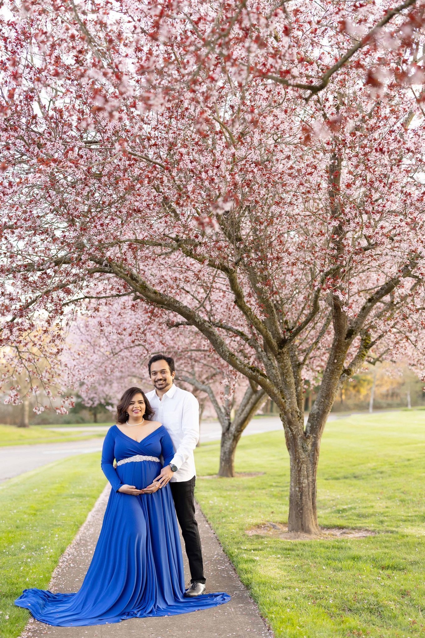 A pregnant woman in a blue dress is standing next to a man under a cherry blossom tree
