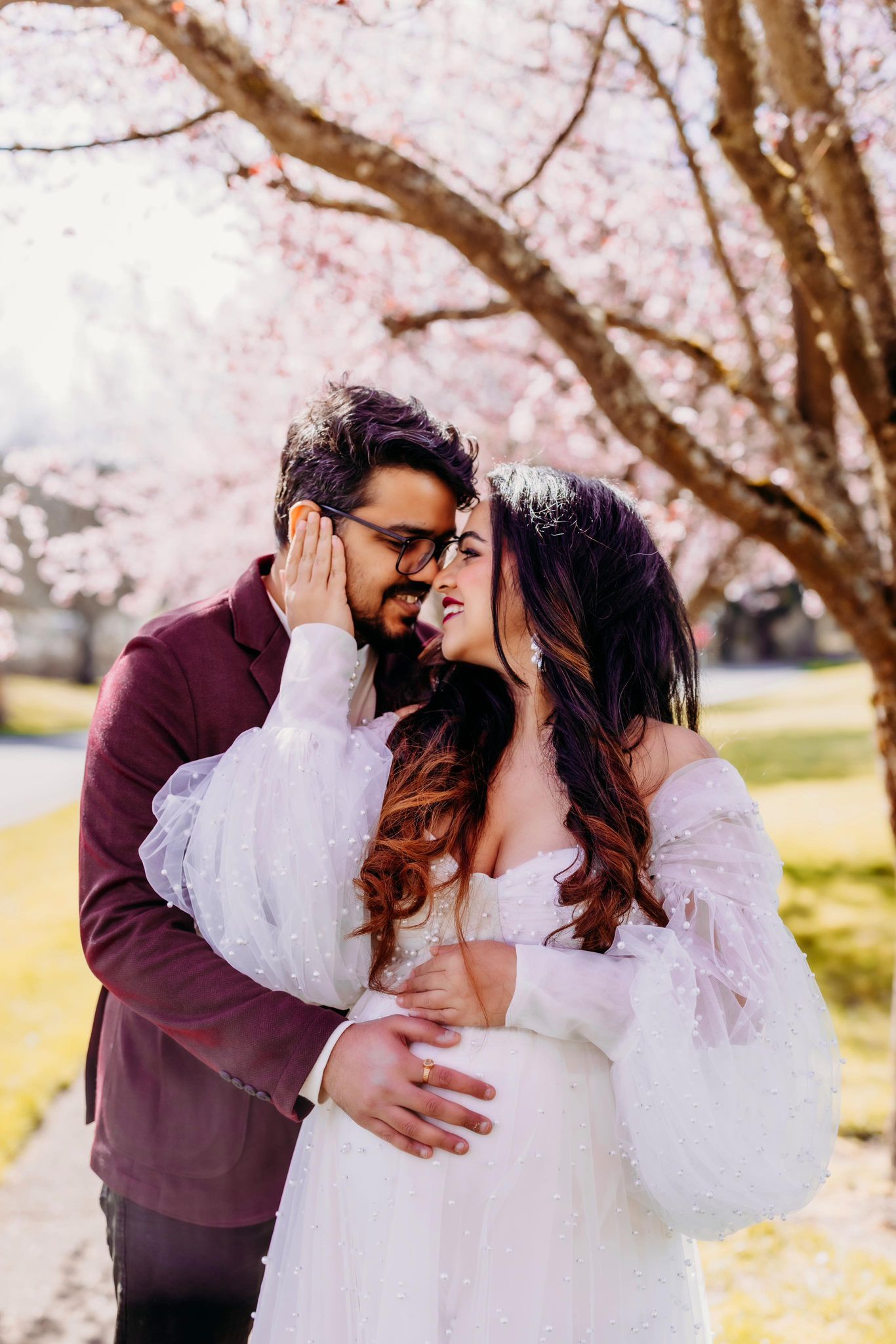 A man and a pregnant woman are posing for a picture under a cherry blossom tree