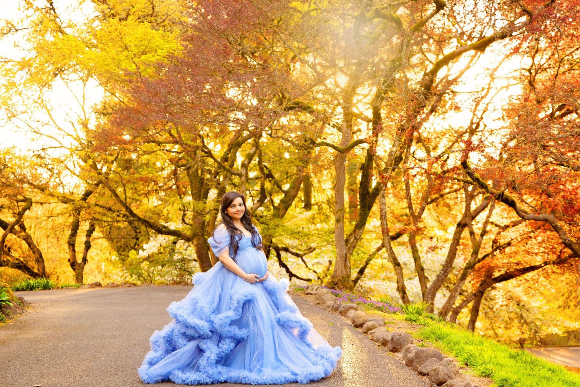 A pregnant woman in a blue gown is standing on a road surrounded by trees