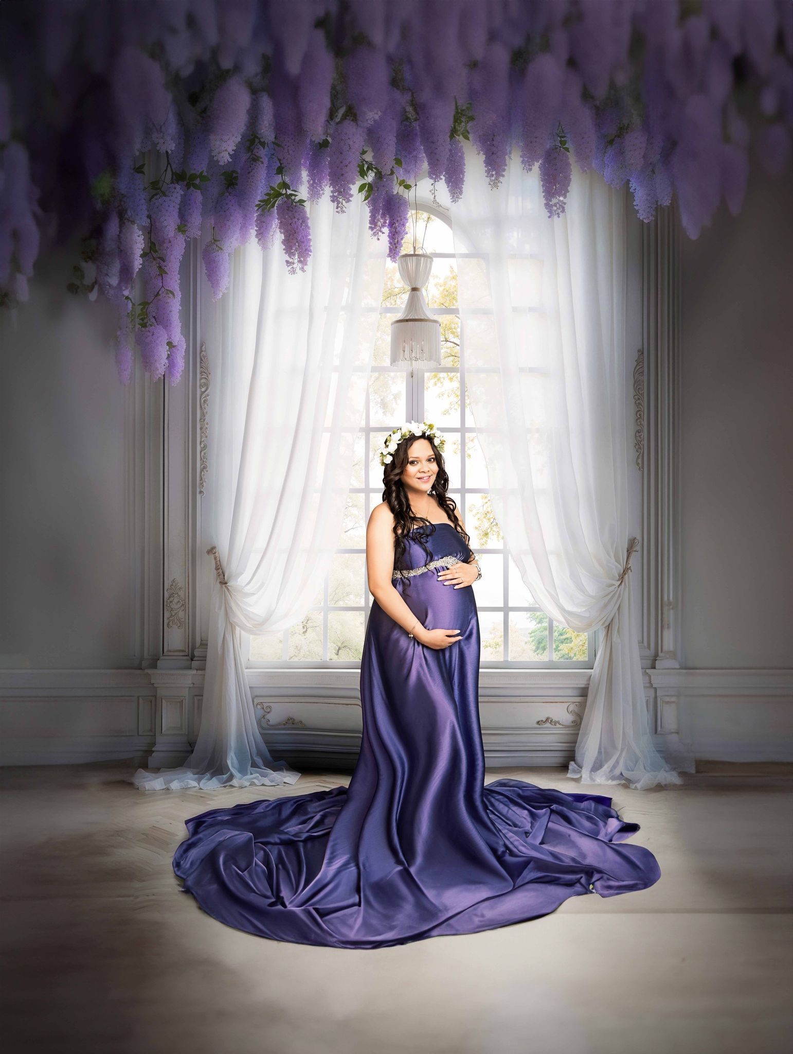 A pregnant woman in a purple dress is standing in front of a window with purple flowers hanging from the ceiling