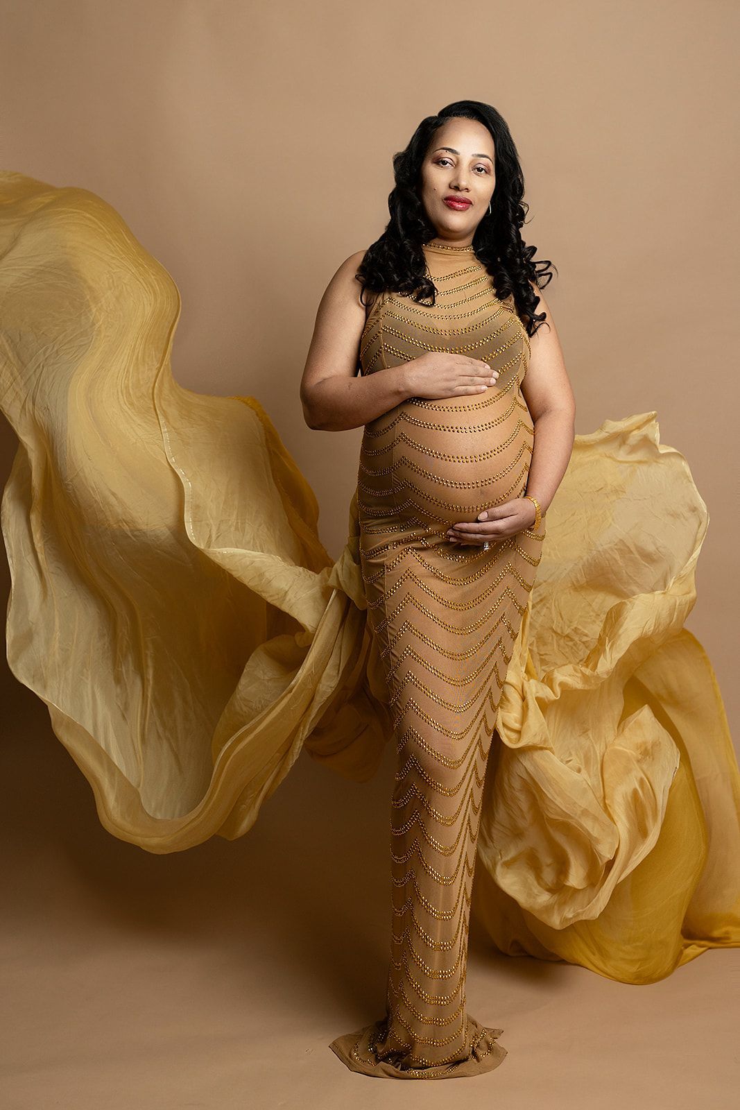 A pregnant woman in a long gold dress is holding her belly.