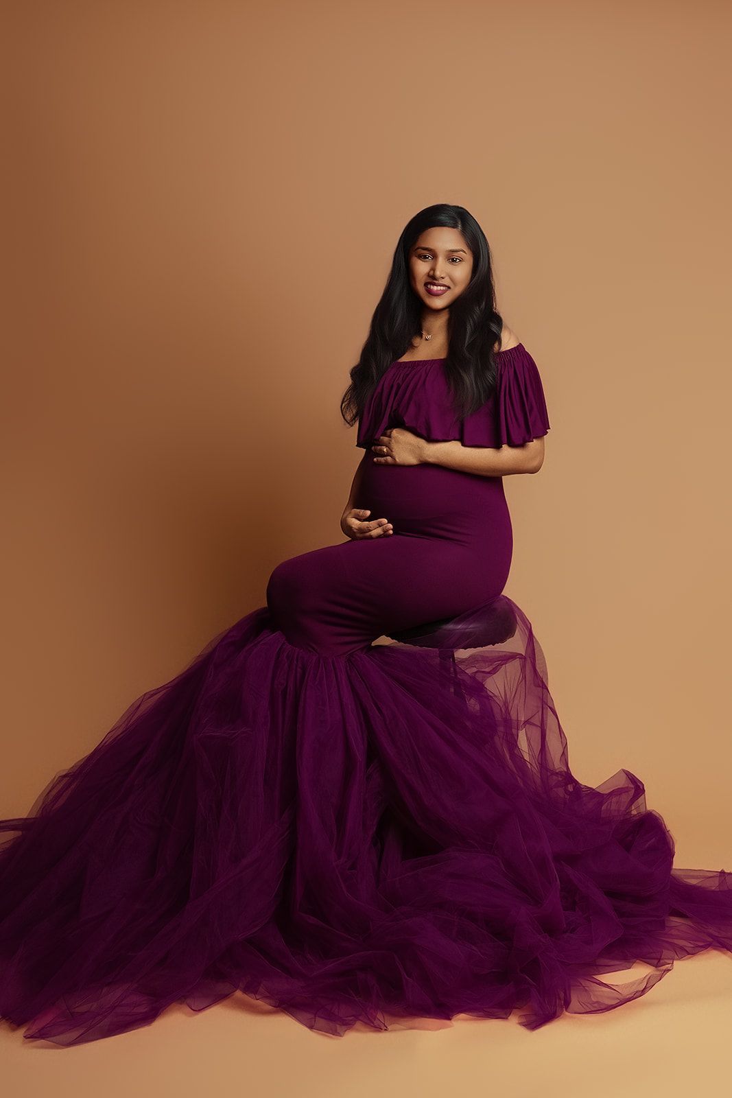 A pregnant woman in a purple dress is sitting on a stool.
