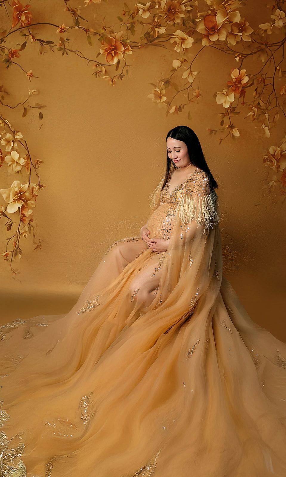 A pregnant woman in a long gold dress is sitting in front of a wall with flowers.