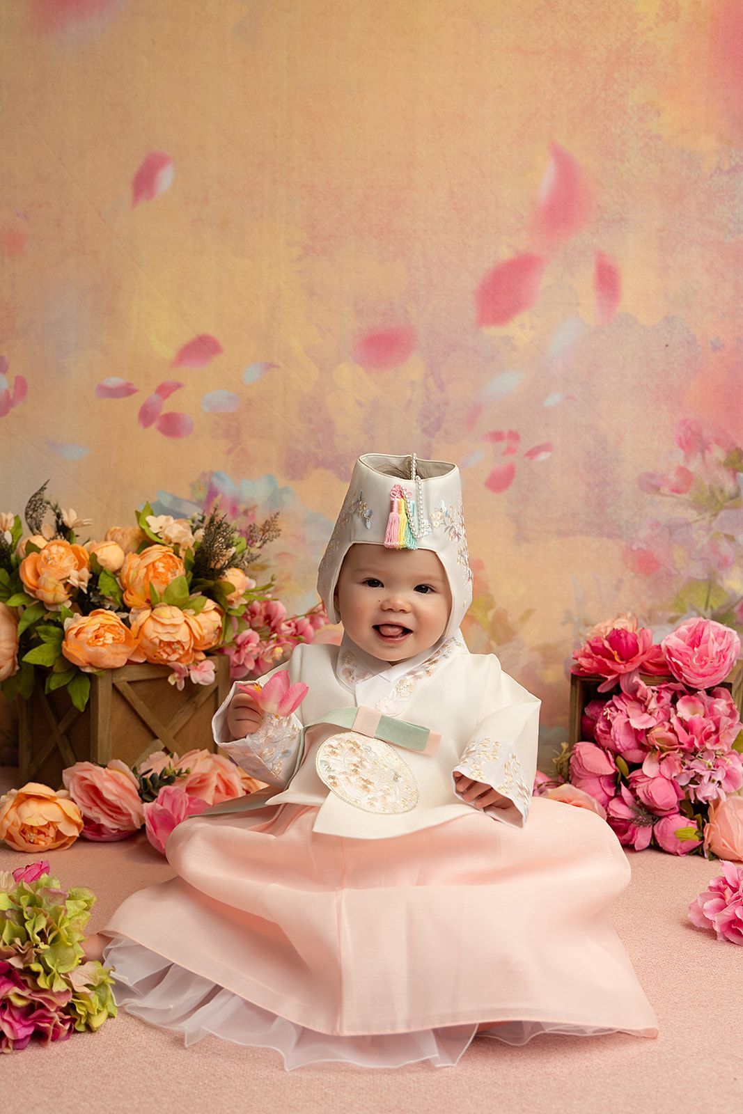 A baby in a pink dress is sitting in front of flowers.
