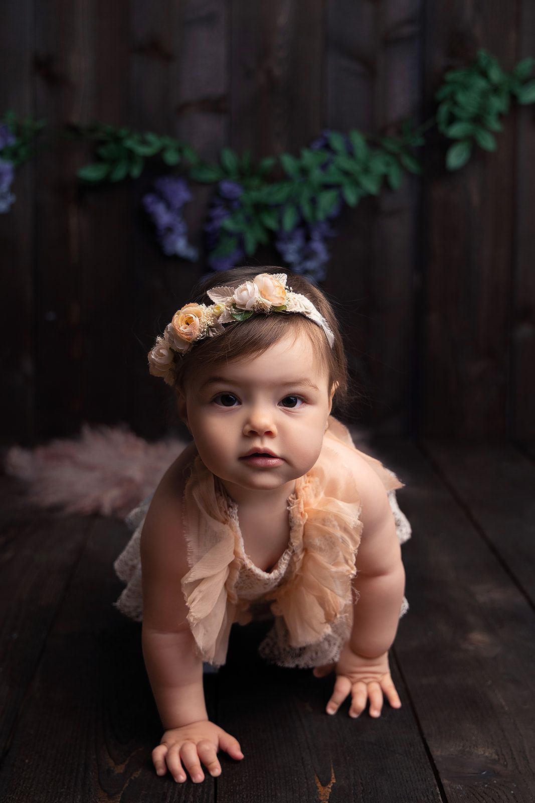 A baby girl wearing a flower crown is crawling on a wooden floor.