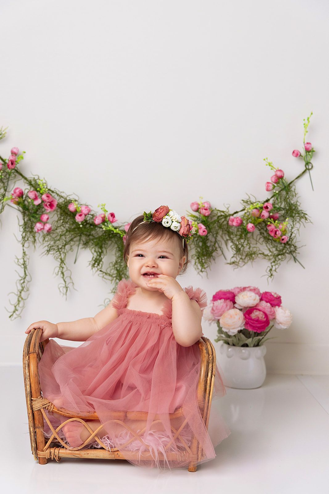 A baby girl is sitting in a wicker basket with flowers in the background.