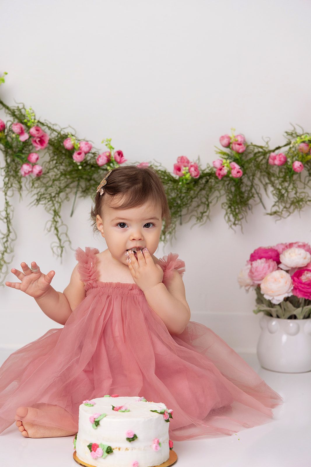 A baby girl in a pink dress is sitting next to a cake.