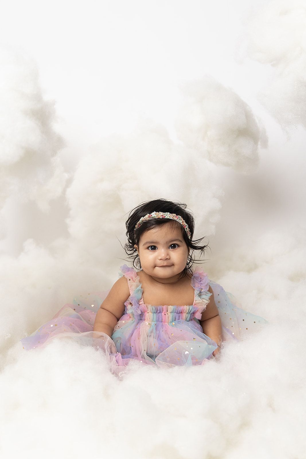 A baby girl in a rainbow dress is sitting in the clouds