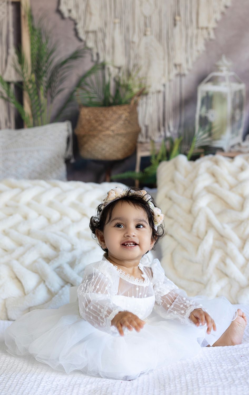 A baby girl in a white dress is sitting on a bed