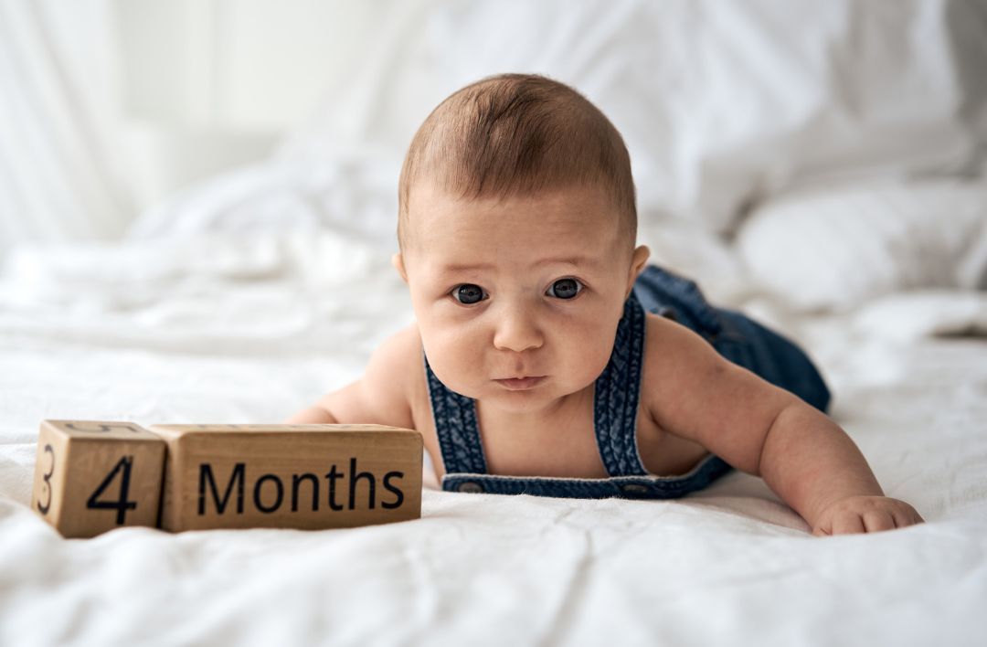 A baby is laying on a bed next to a wooden block that says