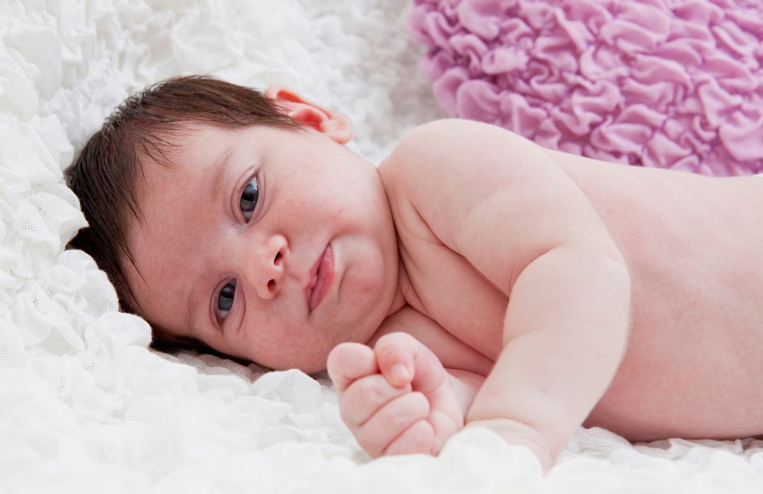 A baby is laying on a white blanket and looking at the camera