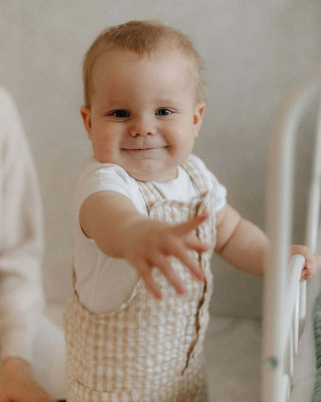 A baby in overalls is standing in a crib and smiling