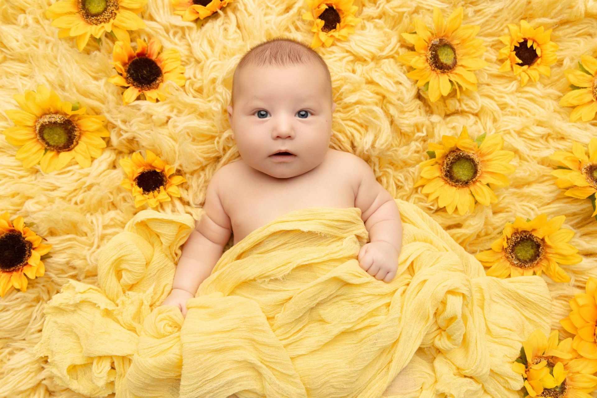A baby is wrapped in a yellow blanket surrounded by sunflowers.