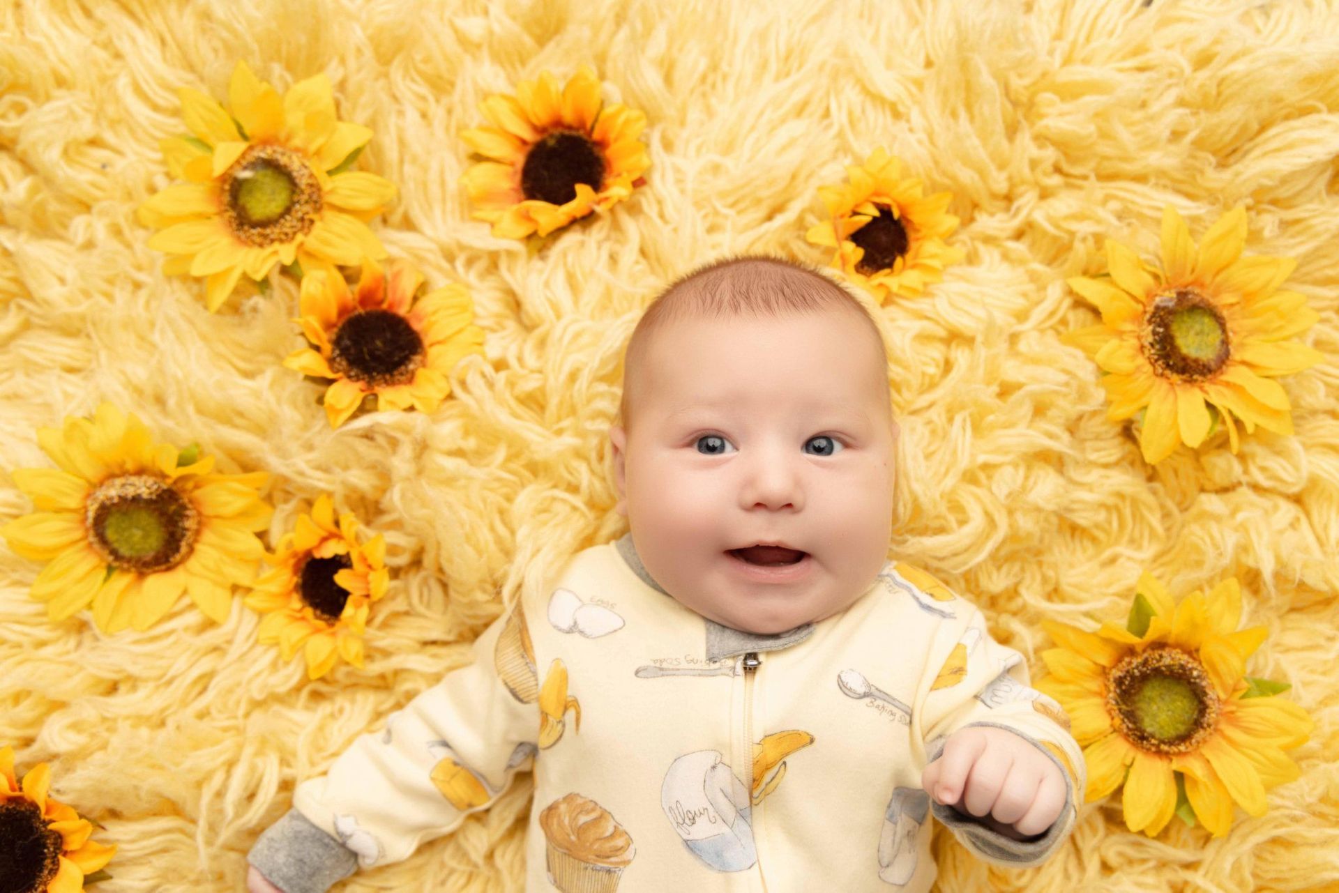 A baby is laying on a yellow blanket surrounded by sunflowers.