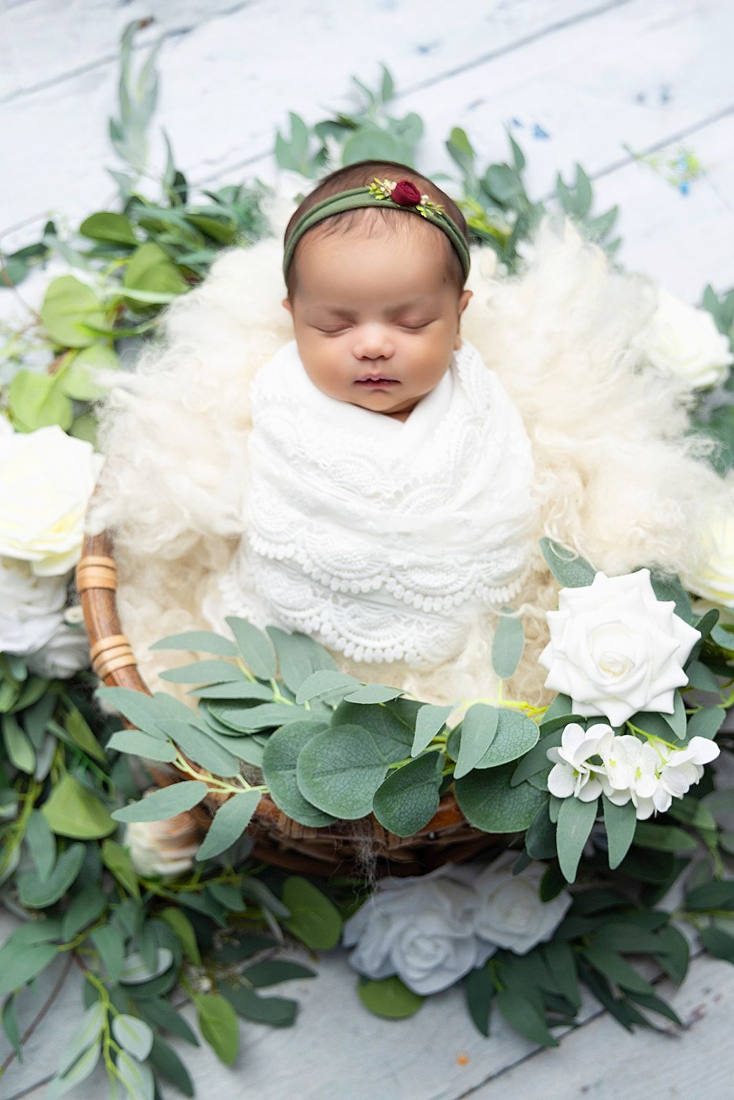 A newborn baby is wrapped in a white blanket and laying in a basket surrounded by flowers.
