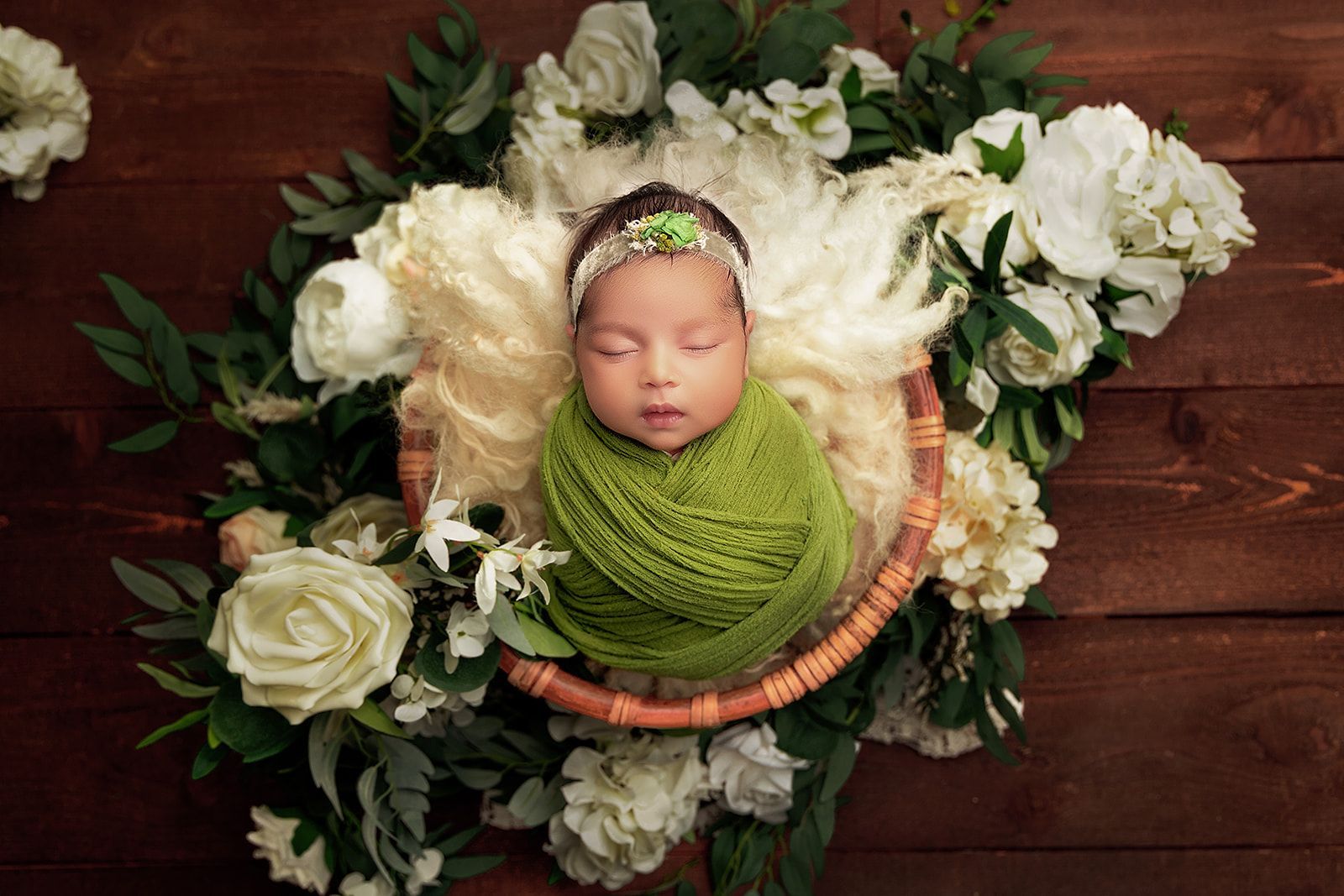 A newborn baby wrapped in a green blanket is sleeping in a basket surrounded by white flowers.
