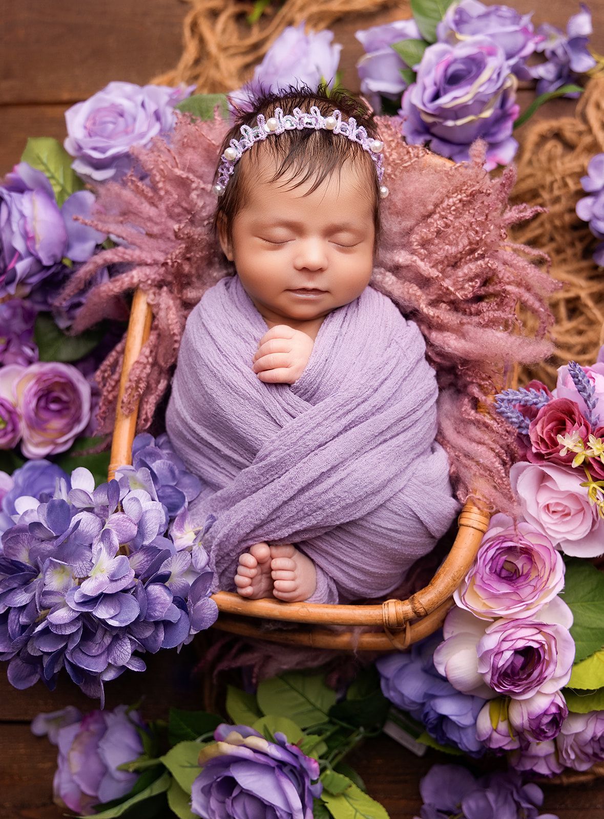 A newborn baby is sleeping in a basket surrounded by purple flowers.