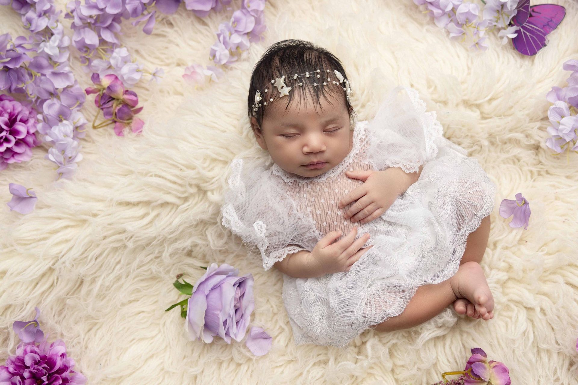A baby in a white dress is laying on a blanket surrounded by purple flowers.