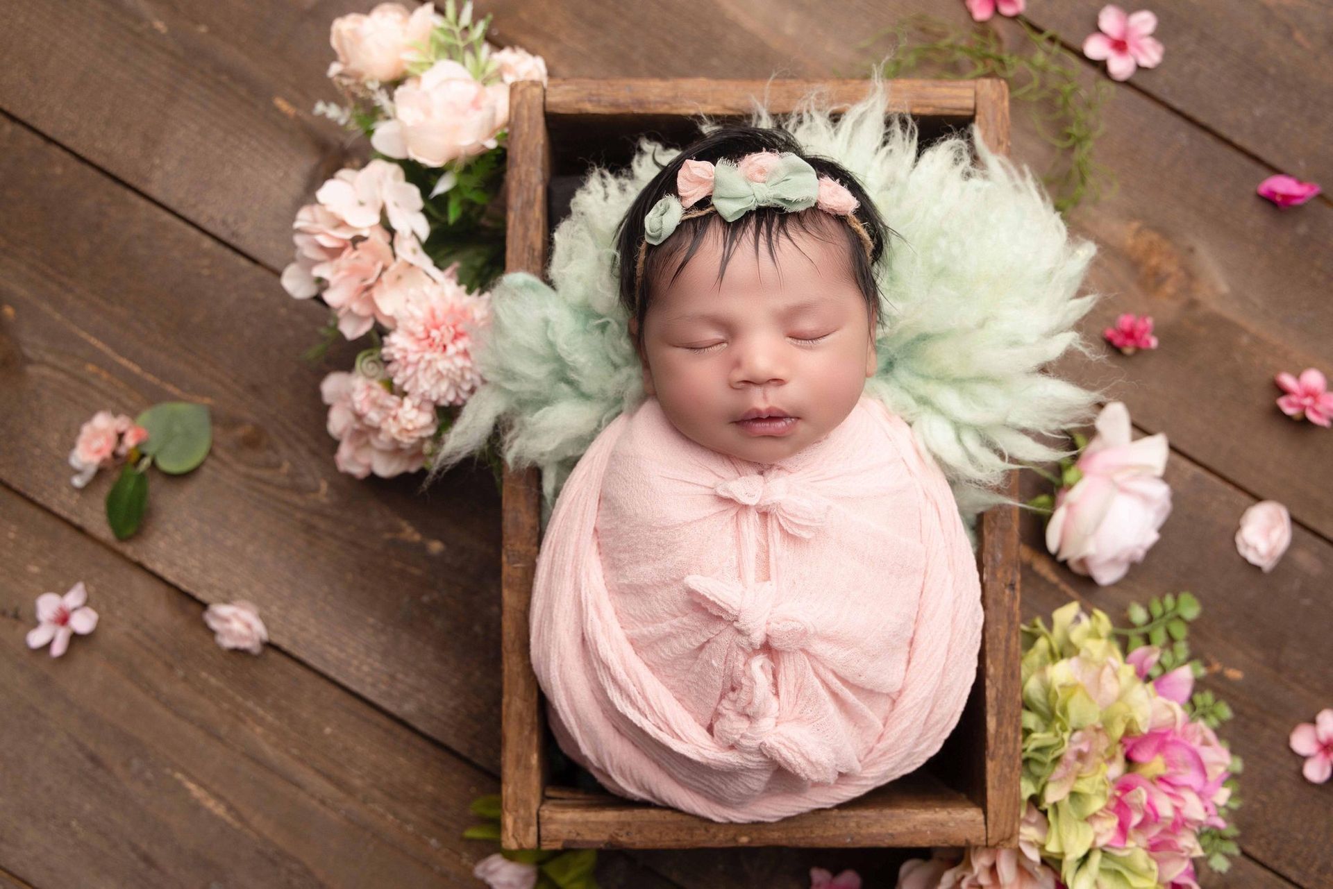A newborn baby wrapped in a pink blanket is sleeping in a wooden box surrounded by flowers.