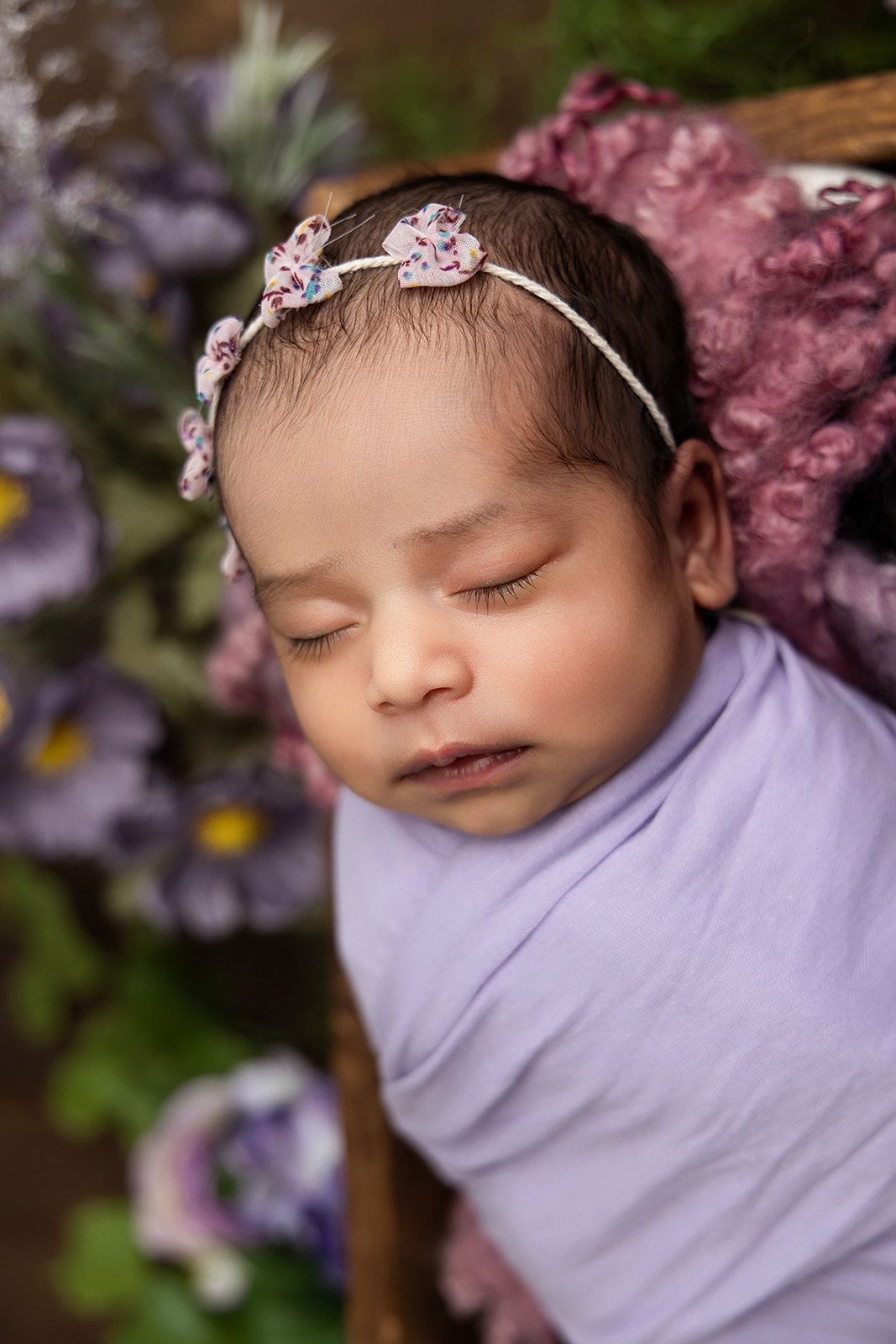 A newborn baby is wrapped in a purple blanket and wearing a headband.