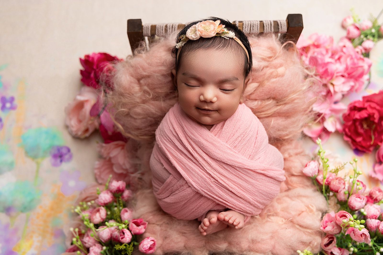 A newborn baby wrapped in a pink blanket is laying on a bed surrounded by flowers.