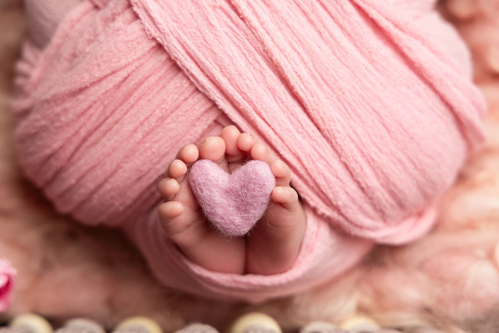A baby wrapped in a pink blanket is holding a pink heart on its feet.