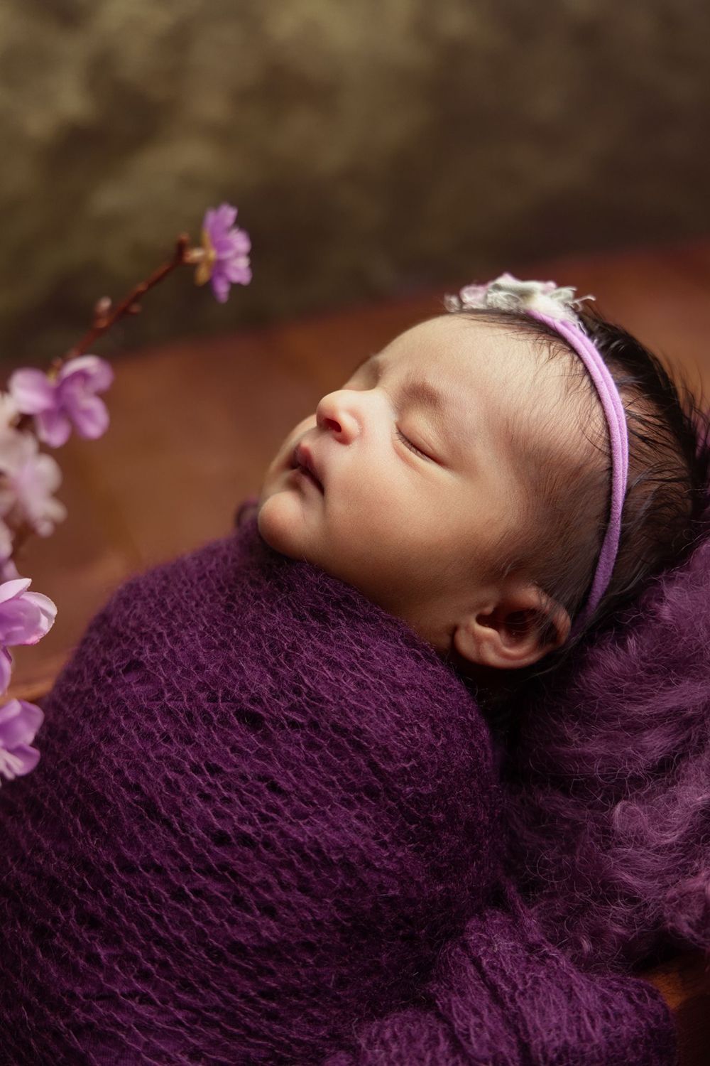 A baby wrapped in a purple blanket is sleeping next to flowers