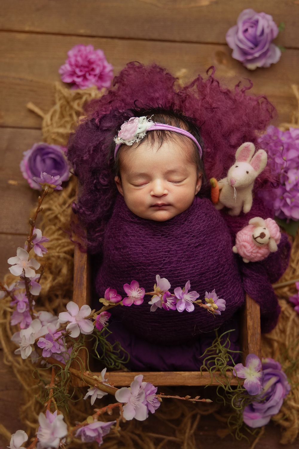 A newborn baby wrapped in a purple blanket is sleeping in a wooden crate surrounded by purple flowers