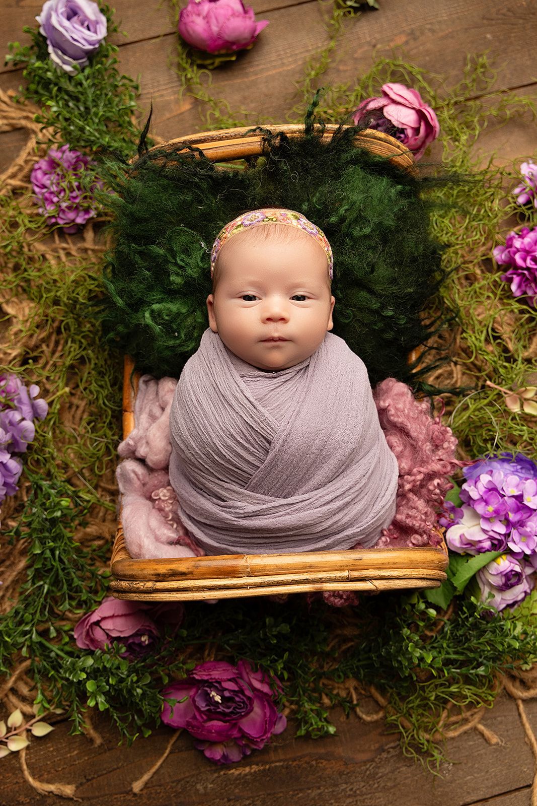 A newborn baby wrapped in a purple blanket is laying in a basket surrounded by flowers