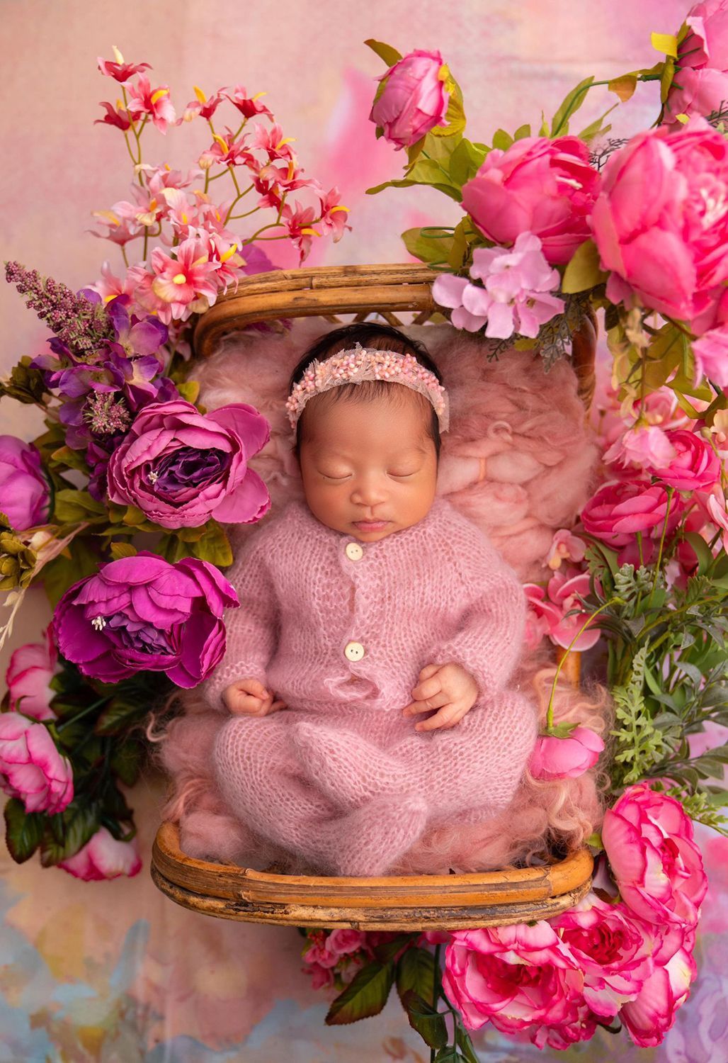 A newborn baby is sleeping in a basket surrounded by pink and purple flowers
