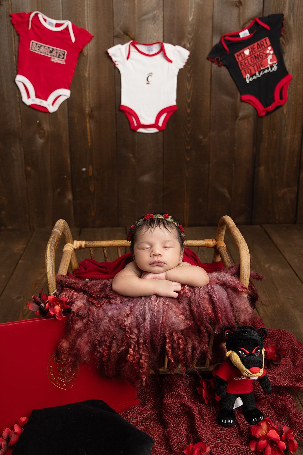 A baby is sleeping in a basket with Cincinnati Bearcats football clothes hanging from the ceiling