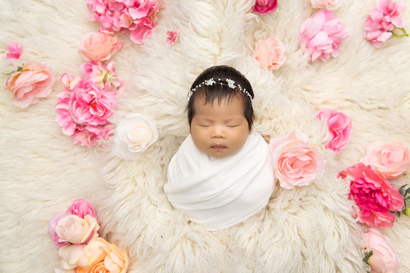 A newborn baby is wrapped in a white blanket and surrounded by pink flowers