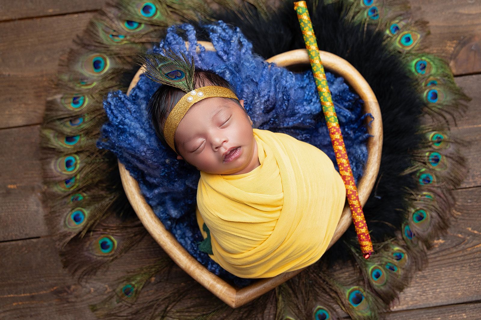 A newborn baby wrapped in a yellow blanket is sleeping in a heart shaped basket surrounded by peacock feathers