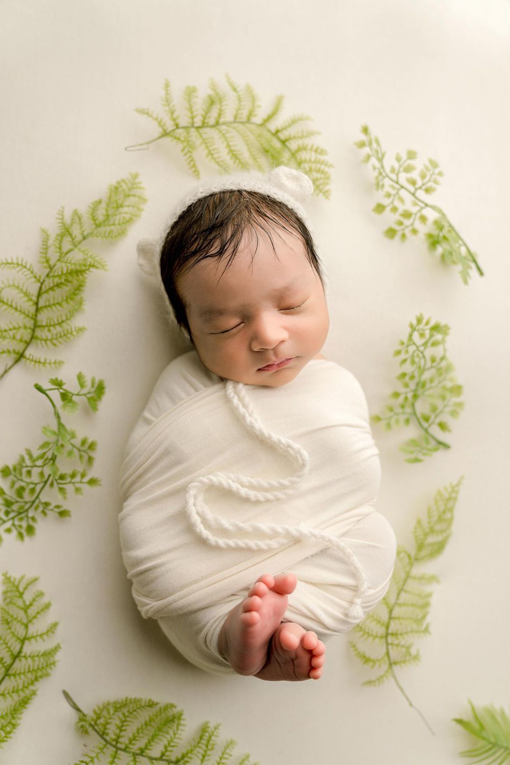 A newborn baby is wrapped in a white blanket and surrounded by green leaves