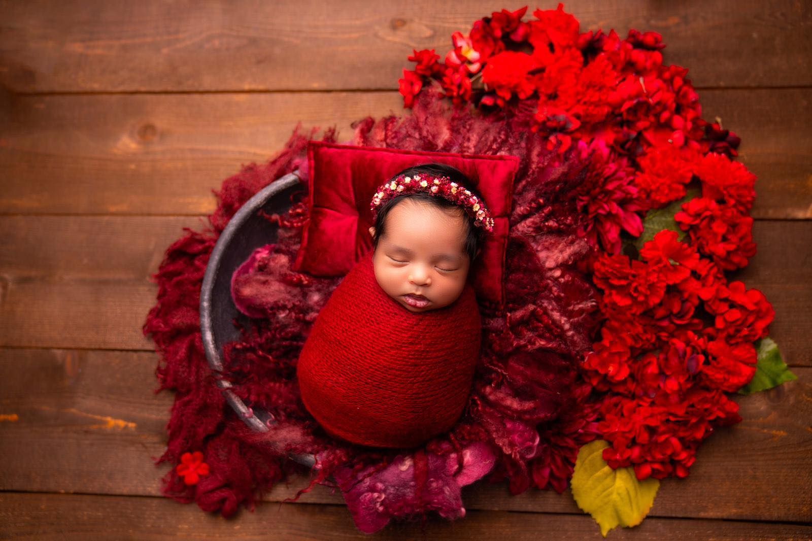 A newborn baby is wrapped in a red blanket and surrounded by red flowers