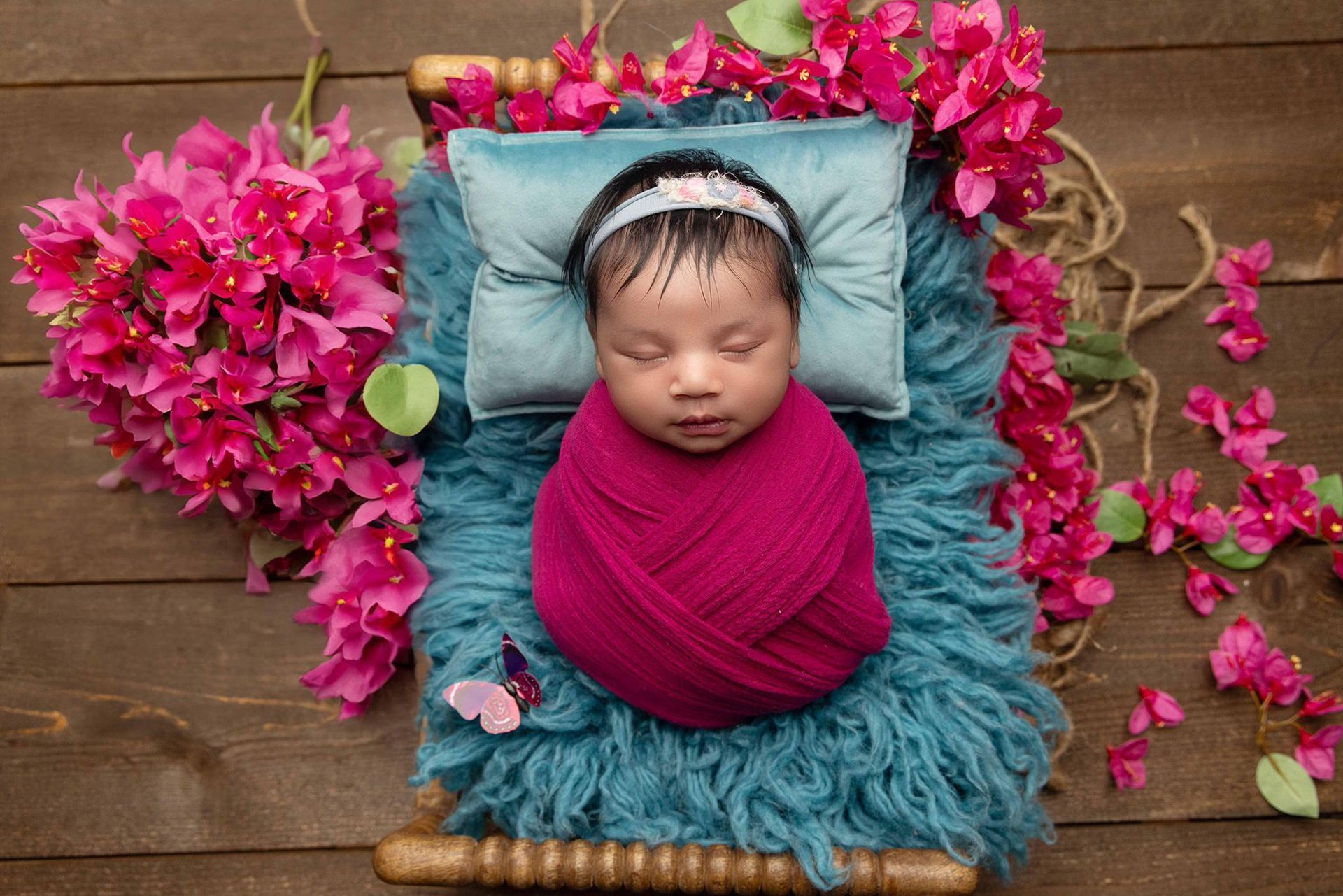 A newborn baby wrapped in a dark pink blanket surrounded by bougainvillea flowers