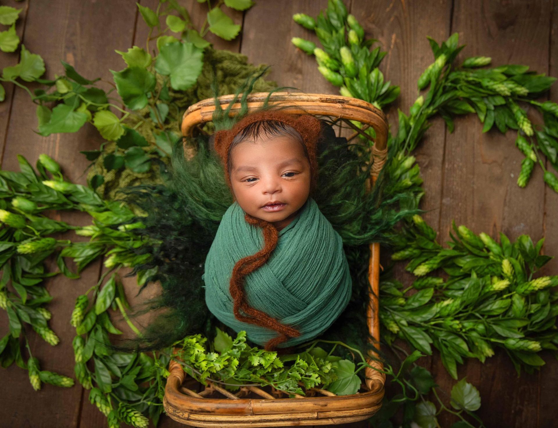A newborn baby wrapped in a green blanket is laying in a wicker basket surrounded by greenery