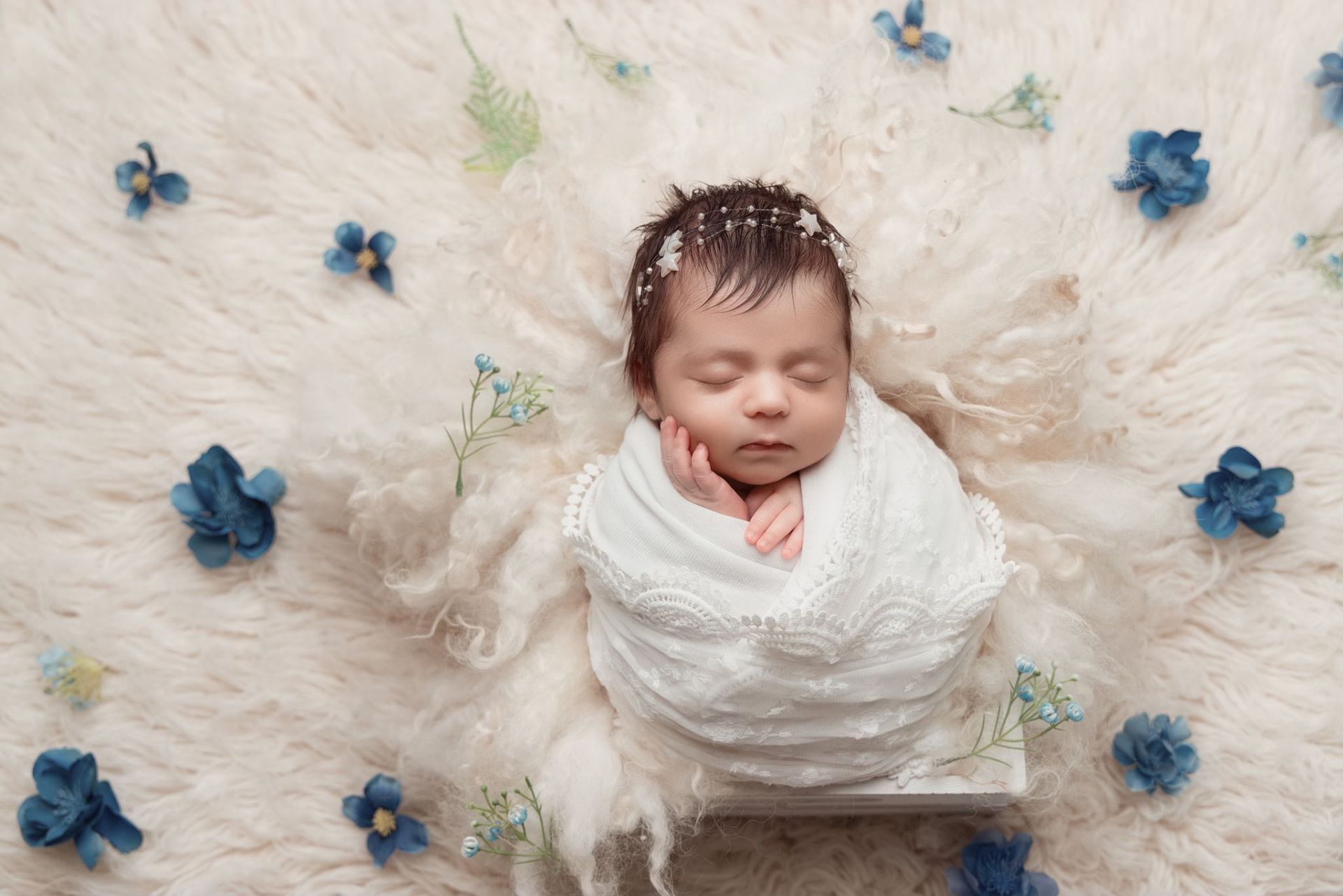 A newborn baby is wrapped in a white blanket and surrounded by blue flowers
