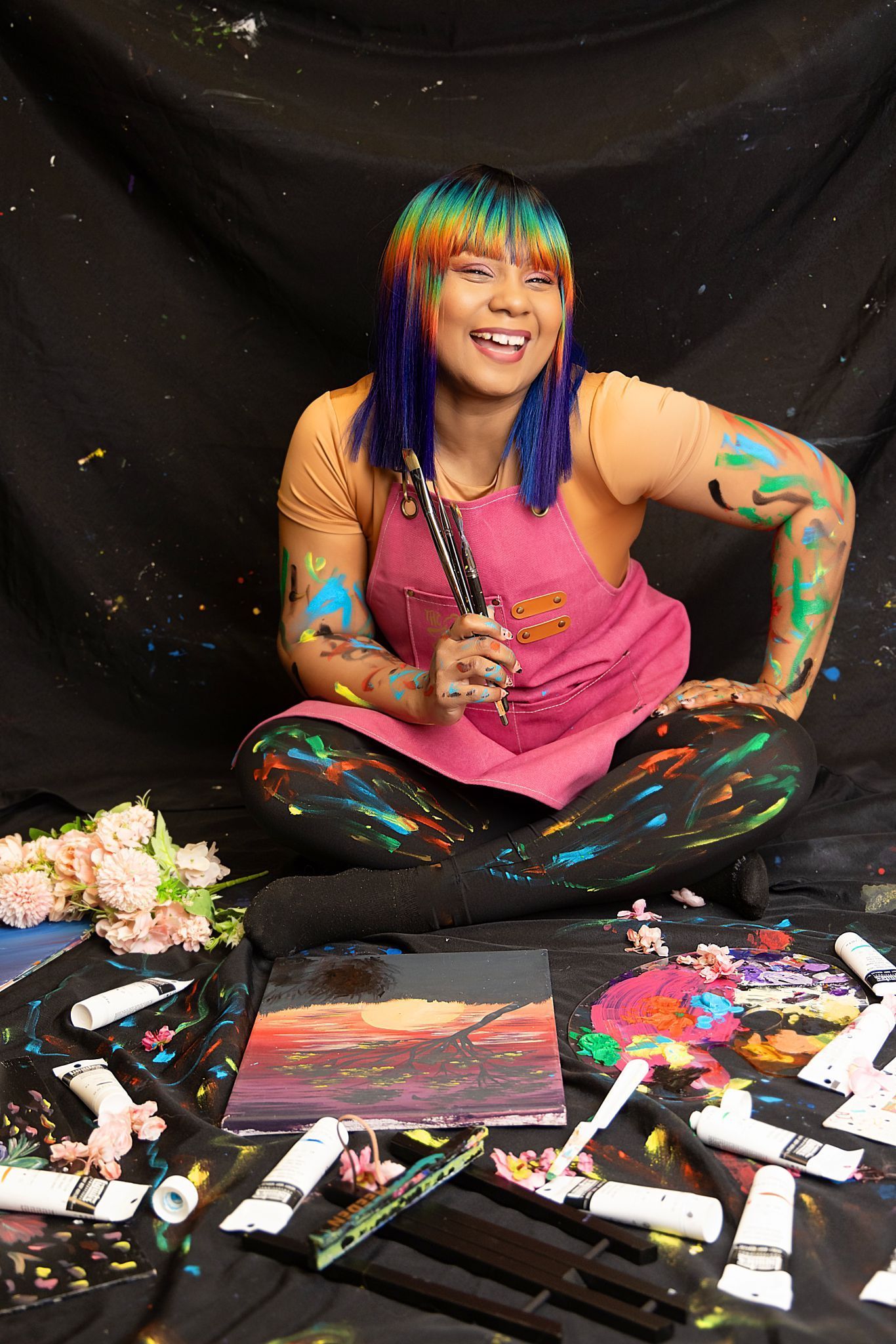 A woman with rainbow hair is sitting on the floor surrounded by paint