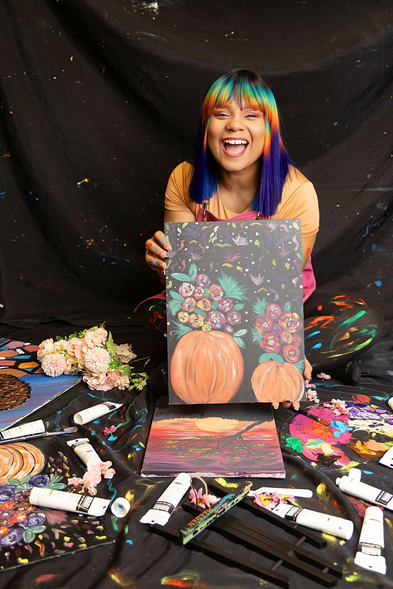A woman with rainbow hair is holding a painting of a pumpkin