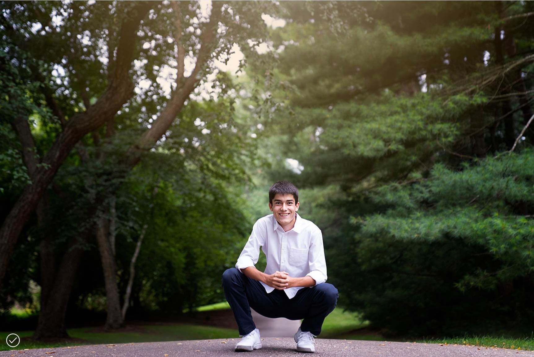 A young man is squatting down in a park with trees in the background