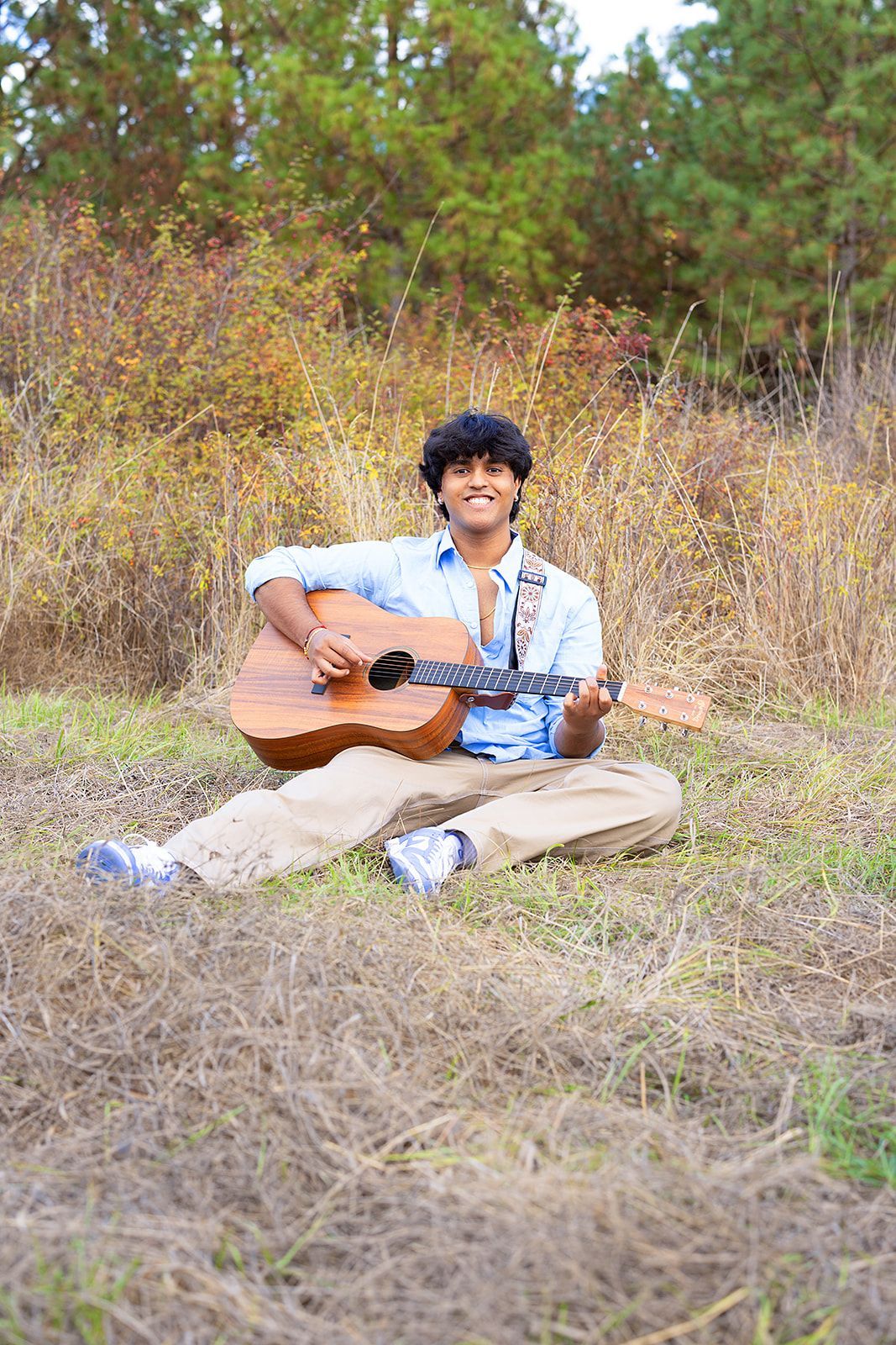 A young man is sitting in a field playing an acoustic guitar.