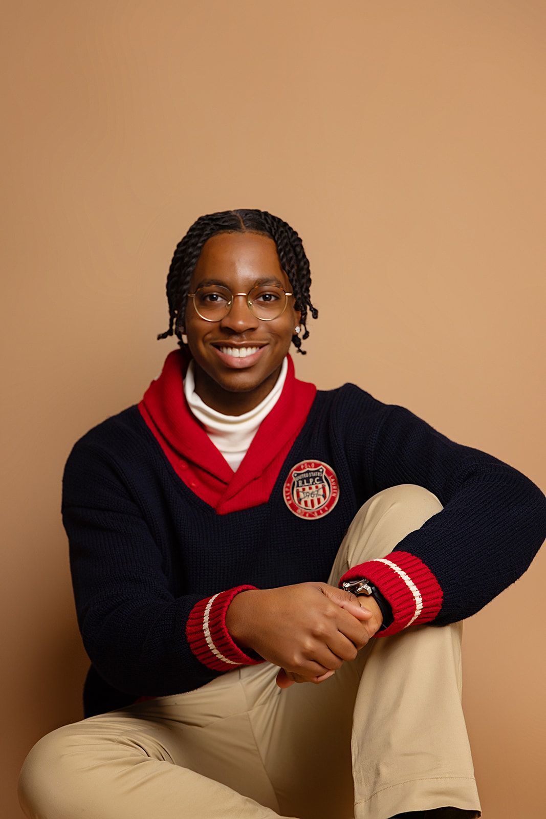 A young man is sitting on the floor wearing a sweater and glasses.